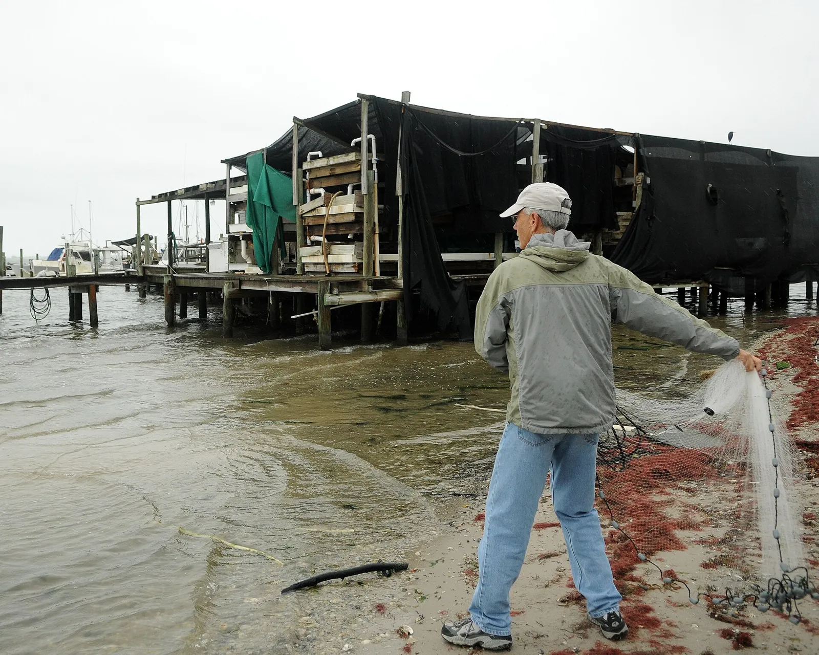 A man in a cap and rain jacket throws a cast net from a sandy shoreline near docks